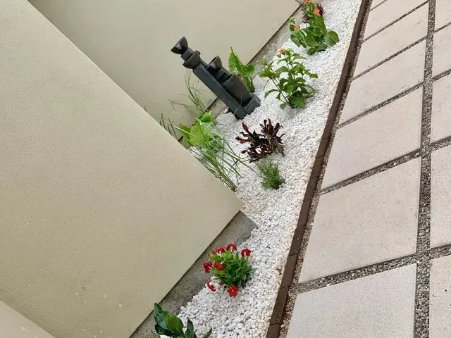 A narrow strip of white gravel between a stucco wall and a run of large pavers, planted through with small red-flowered plants
