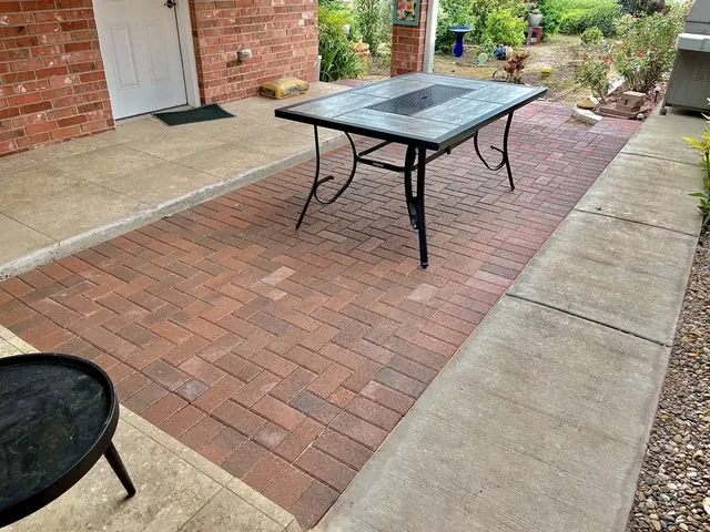 A red and tan paver patio laid in herringbone beside a red brick house, with a patio table on it and a fire bowl in the foreground