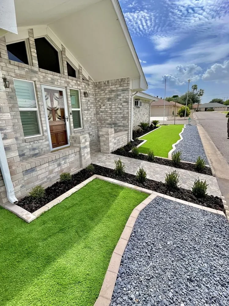 A front yard laid out in panels of bright artificial turf, black rock beds planted with small shrubs and blue-gray gravel, all edged in pale curbing, with a paver walk up to the door of a pale brick house