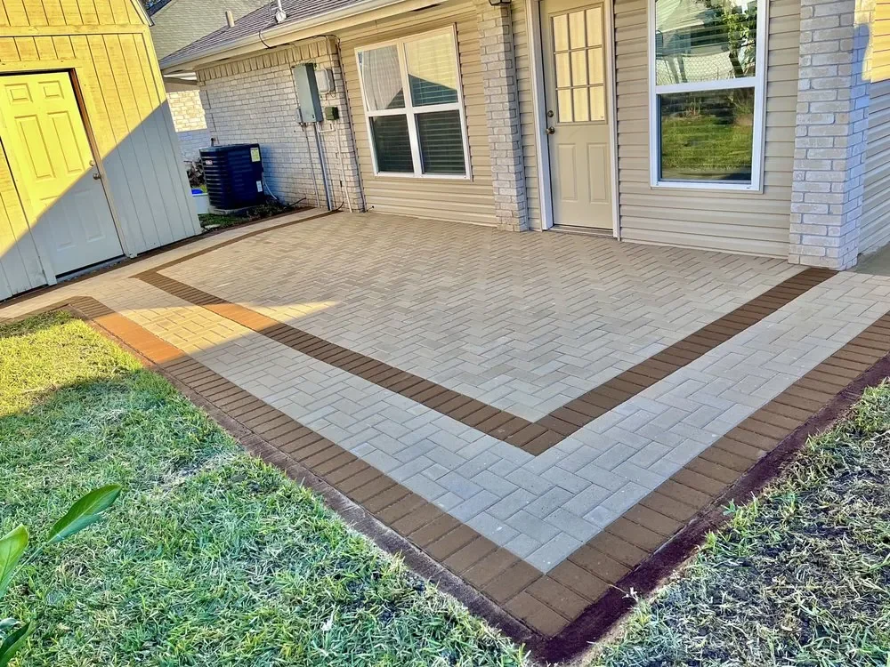 A back patio of pale gray pavers laid in herringbone inside a dark brown border band, running up to the back of a brick house with lawn across the foreground